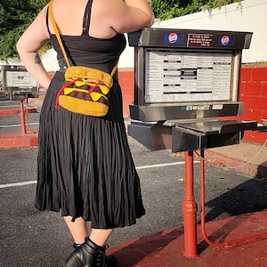 May include: A person wearing a black skirt and a black tank top with a crocheted hamburger purse. The purse is a bright yellow with red and green accents. The person is standing in front of a red and white building with a menu board.