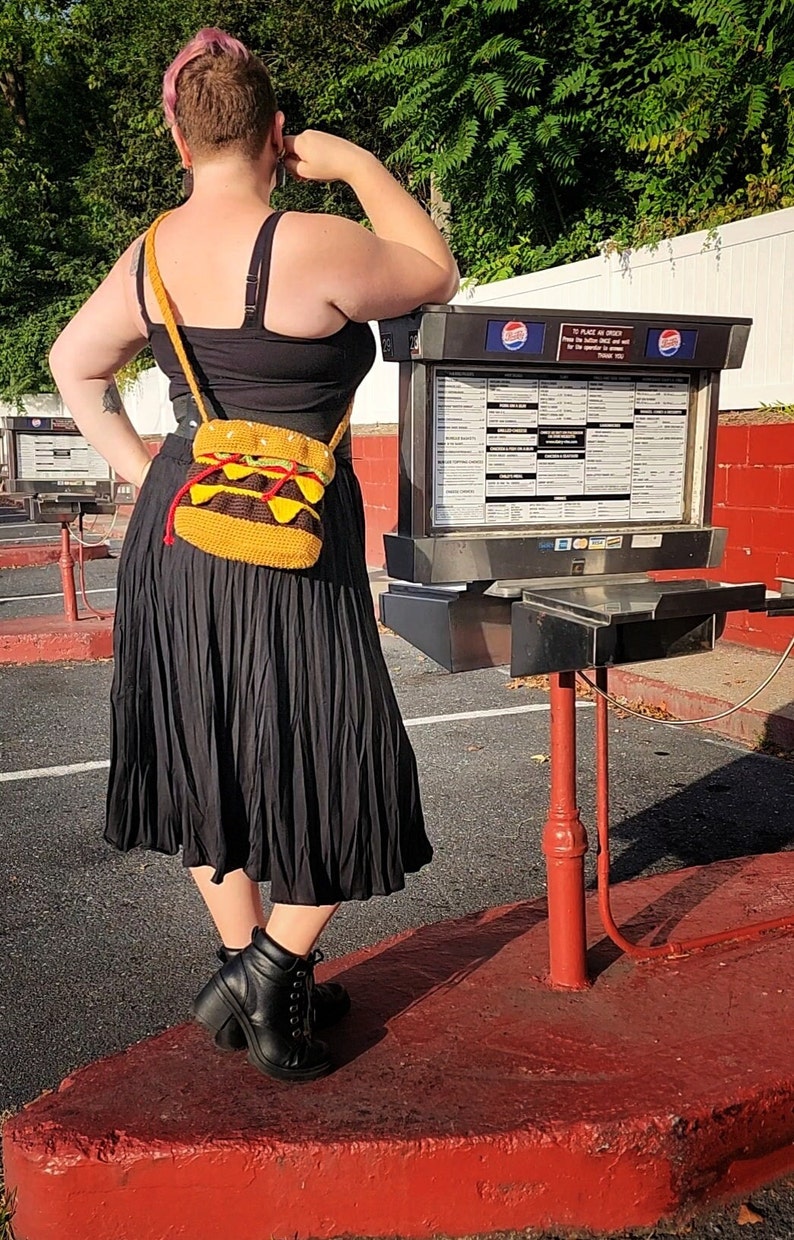 May include: A person wearing a black skirt and a black tank top with a crocheted hamburger purse. The purse is a bright yellow with red and green accents. The person is standing in front of a red and white building with a menu board.