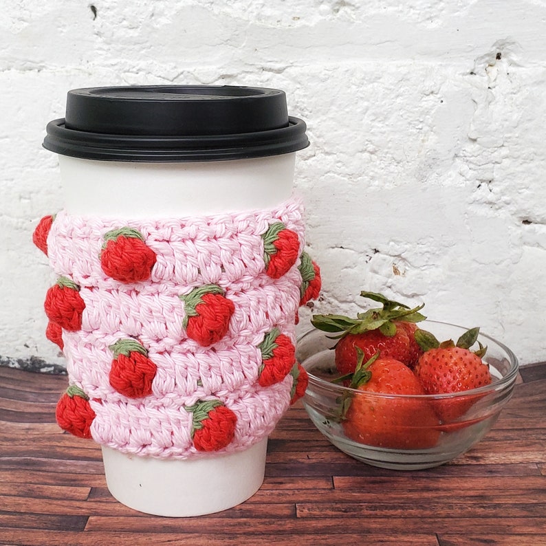 May include: A pink crocheted coffee cup cozy with red strawberry embellishments. The cozy is on a white to-go coffee cup. A bowl of fresh strawberries is in the background.