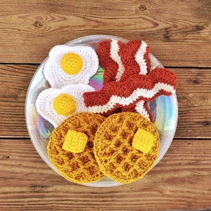 K&ouml;nnte beinhalten: Ein geh&auml;keltes Fr&uuml;hst&uuml;cksset mit zwei gelben und braunen Waffeln, zwei wei&szlig;en Eiern mit gelben Dottern und zwei Streifen rot-wei&szlig;em Speck auf einem wei&szlig;en Teller mit Regenbogenmuster.