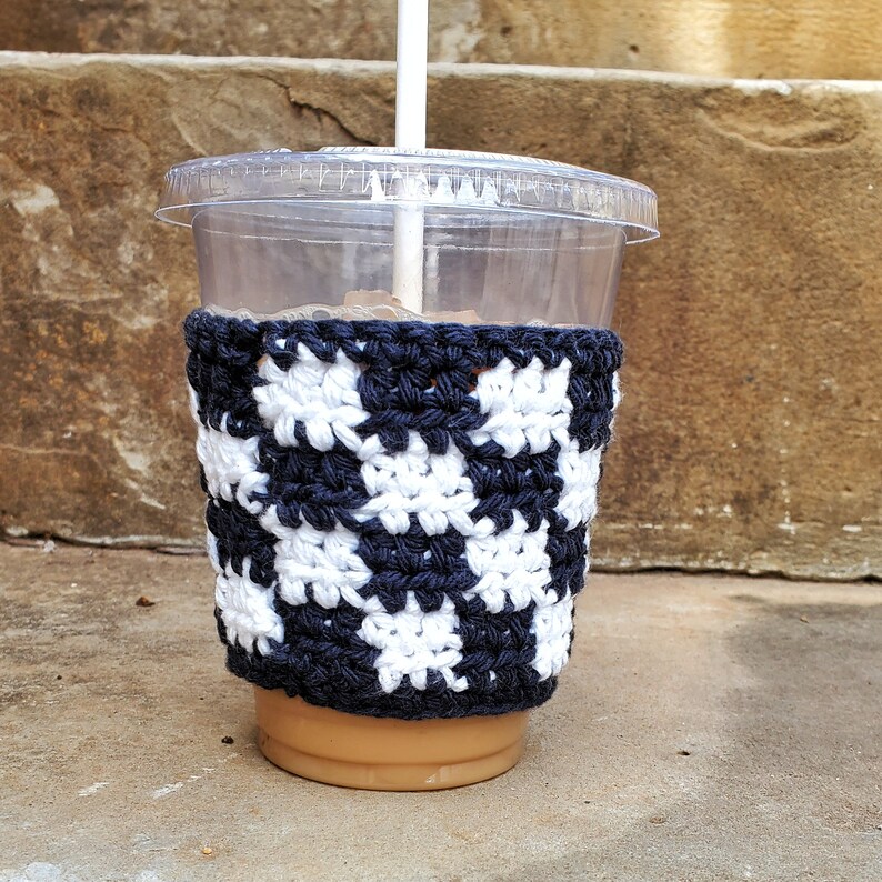 May include: A clear plastic cup with a crocheted beverage cozy. The cozy is black and white with a geometric pattern. The cup is filled with a brown liquid.