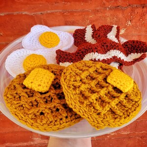 K&ouml;nnte beinhalten: Ein geh&auml;keltes Fr&uuml;hst&uuml;cksessen-Set mit zwei gelben Waffeln, zwei wei&szlig;en Eiern und einem rot-wei&szlig;en geh&auml;kelten Speckstreifen. Das Set wird auf einem wei&szlig;en Teller pr&auml;sentiert.