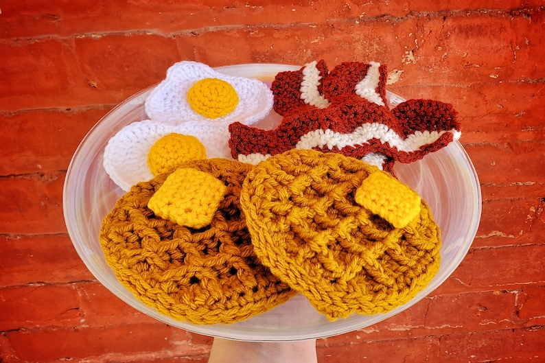K&ouml;nnte beinhalten: Ein geh&auml;keltes Fr&uuml;hst&uuml;cksessen-Set mit zwei gelben Waffeln, zwei wei&szlig;en Eiern und einem rot-wei&szlig;en geh&auml;kelten Speckstreifen. Das Set wird auf einem wei&szlig;en Teller pr&auml;sentiert.