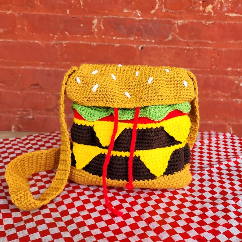 May include: A crocheted hamburger purse with a yellow bun, green lettuce, red tomato, and brown patty. The purse has a long strap and is made of yarn.