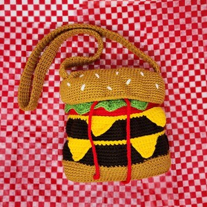 May include: A crocheted hamburger purse with a brown bun, green lettuce, red tomato, yellow cheese, and black patty. The purse has a long brown strap.