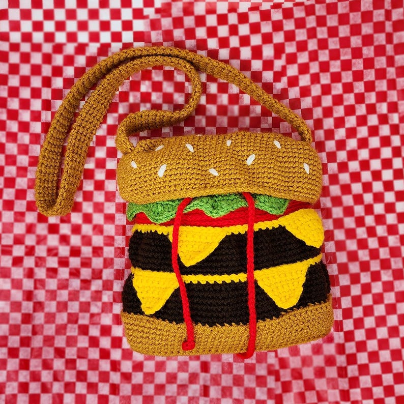 May include: A crocheted hamburger purse with a brown bun, green lettuce, red tomato, yellow cheese, and black patty. The purse has a long brown strap.