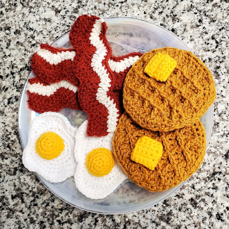 K&ouml;nnte beinhalten: Ein geh&auml;keltes Fr&uuml;hst&uuml;cksset mit zwei gelben und braunen Pfannkuchen, zwei wei&szlig;en Eiern und drei roten und wei&szlig;en Speckscheiben auf einem wei&szlig;en Teller.