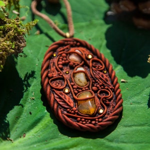 Può includere: Un ciondolo in argilla marrone con un design dettagliato che presenta volute, foglie e piccole perline. Il ciondolo ha tre pietre incastonate, una bianca, una arancione e una ambra.