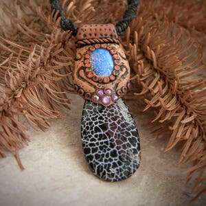 Può includere: Un ciondolo in ceramica marrone e nero con una pietra di opale blu e strass. Il ciondolo è attaccato a un cordoncino nero con una perla bianca.