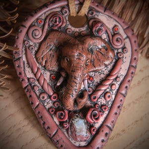 Può includere: Un ciondolo in argilla rosa con una scultura di elefante dettagliata, circondato da motivi floreali rossi e bianchi. Il ciondolo ha un cordino in pelle e accenti di pietre preziose rosse.