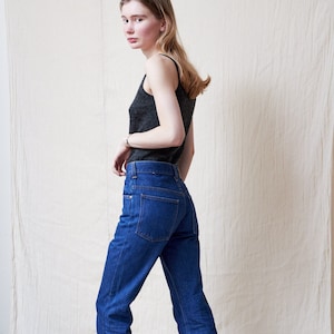 May include: A woman wearing a gray tank top and blue jeans. She is standing in front of a white background.