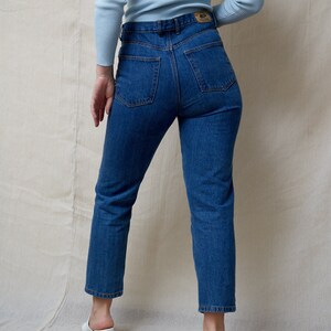 May include: A person wearing a light blue long-sleeved shirt and dark blue jeans. The jeans have a high waist and a straight leg.