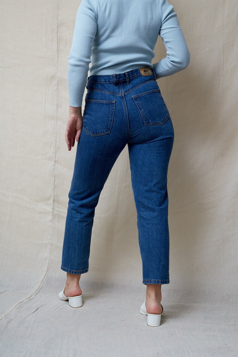 May include: A person wearing a light blue long-sleeved shirt and dark blue jeans. The jeans have a high waist and a straight leg.