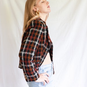 May include: A woman wearing a cropped, brown, black, and white plaid jacket with gold hoop earrings. She is wearing light blue jeans and has her hand on her hip.