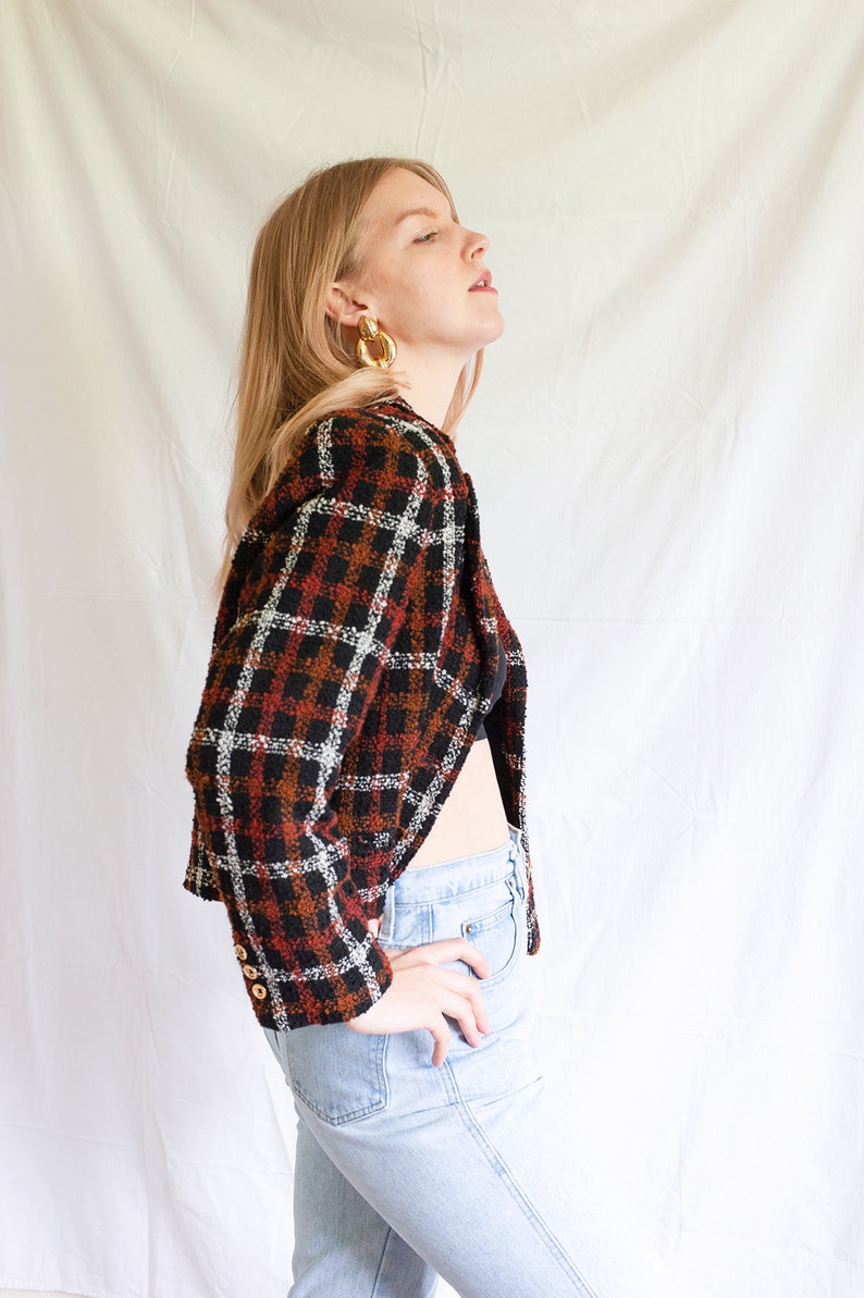 May include: A woman wearing a cropped, brown, black, and white plaid jacket with gold hoop earrings. She is wearing light blue jeans and has her hand on her hip.