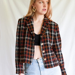 May include: A cropped black, red, and white plaid blazer with gold buttons. The blazer is made of a textured fabric and has a relaxed fit.