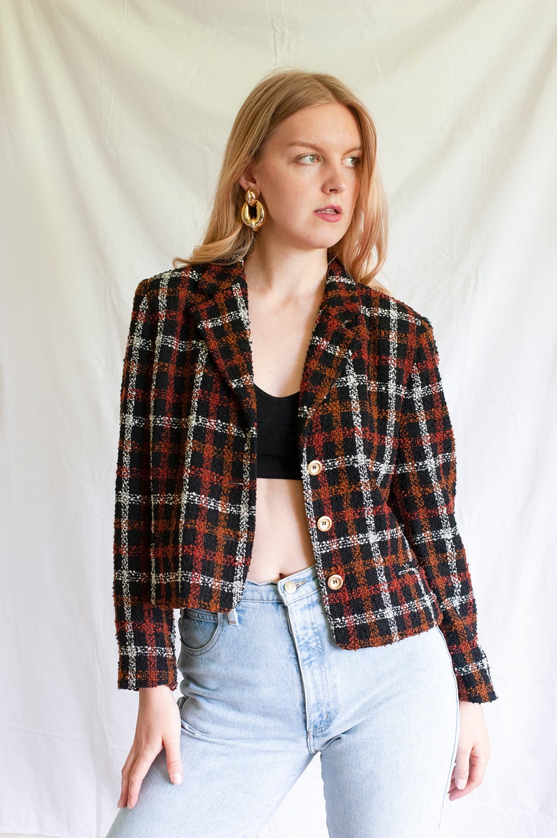 May include: A cropped black, red, and white plaid blazer with gold buttons. The blazer is made of a textured fabric and has a relaxed fit.