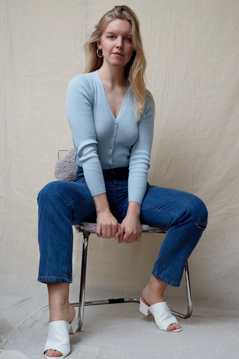 May include: A woman wearing a light blue ribbed cardigan with a v-neck and button closure, blue jeans, and white heeled mules. She is sitting on a metal chair with her hands clasped in her lap.