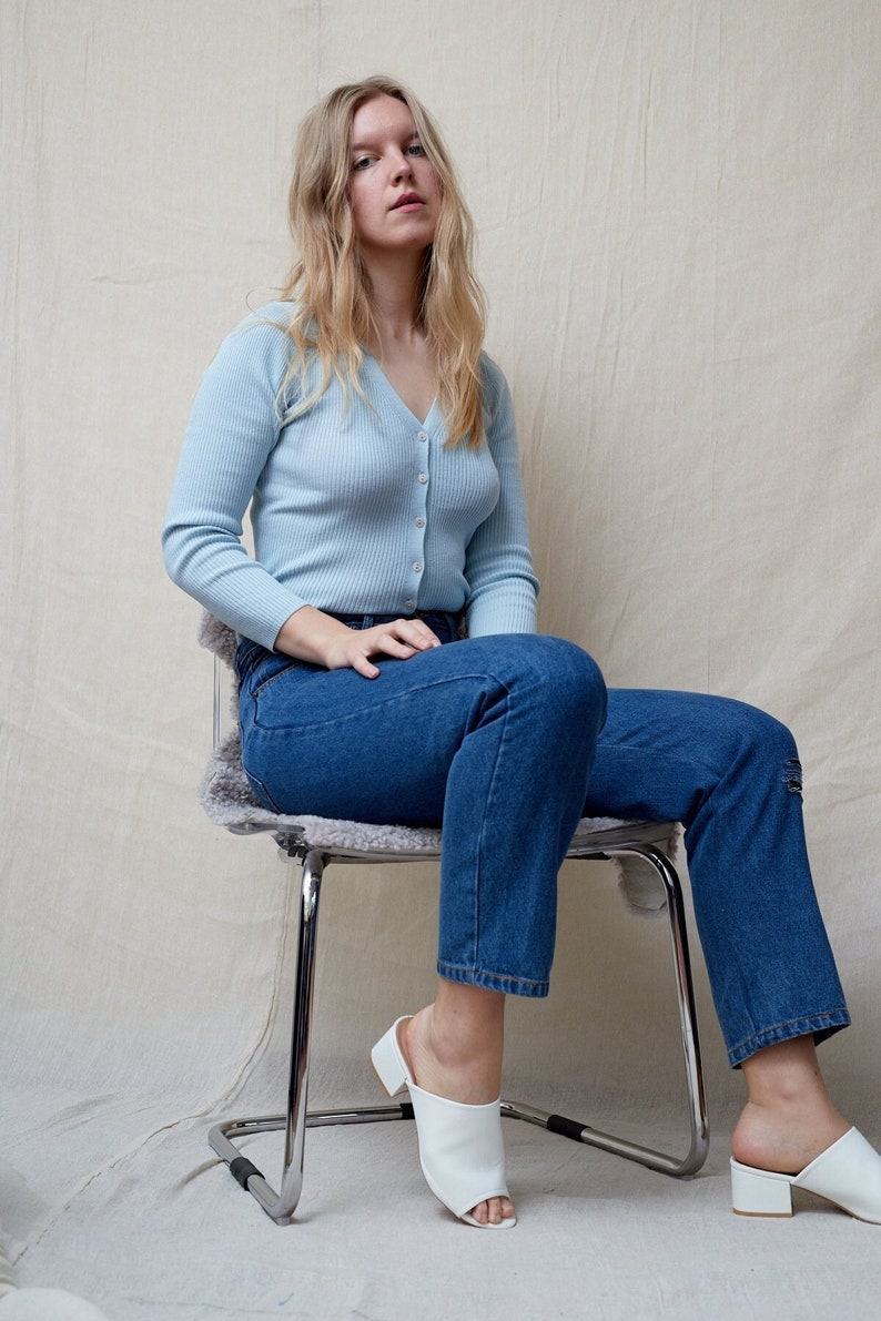 May include: A woman wearing a light blue cardigan and blue jeans sits on a metal chair with a furry seat cushion. She is wearing white open-toe heeled shoes.