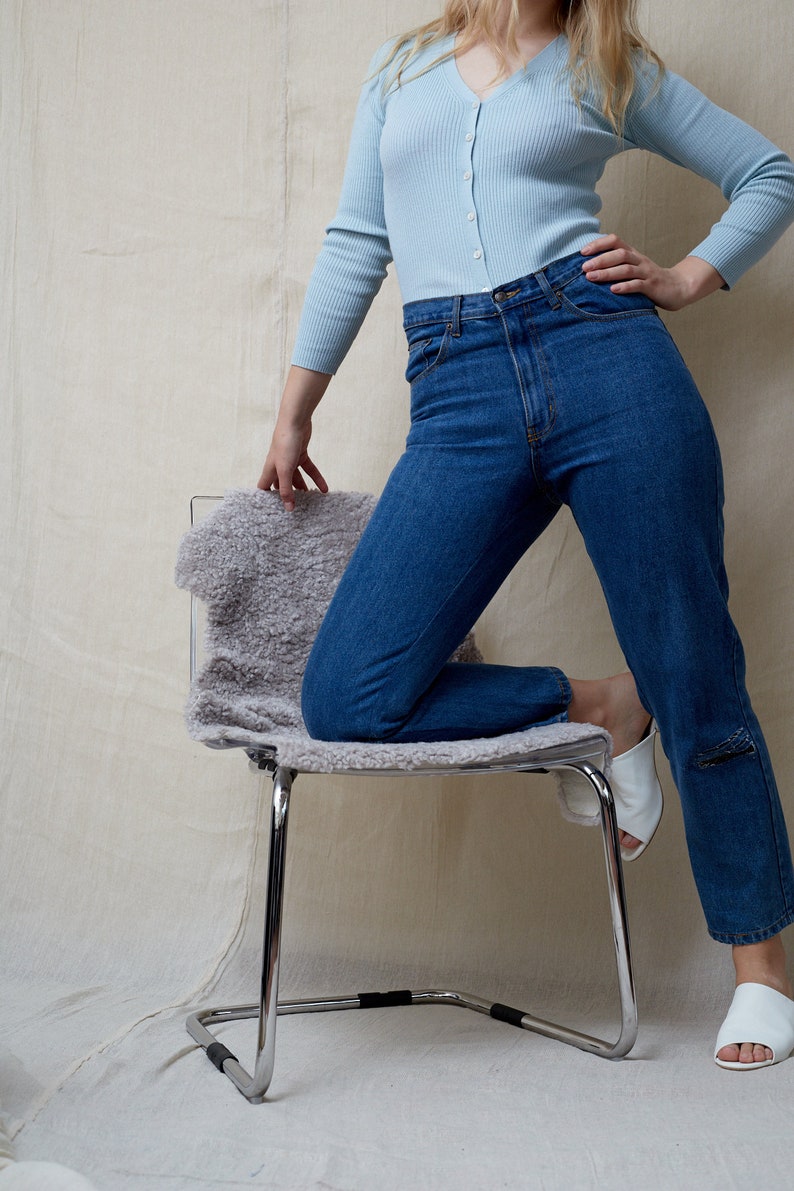 May include: A person wearing a light blue cardigan and blue jeans is kneeling on a chair with a fluffy white cushion. The chair has a chrome frame.