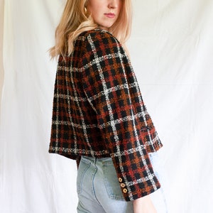 May include: A cropped plaid jacket with a black, white, and orange pattern. The jacket has long sleeves and a button closure. The model is wearing light blue jeans.
