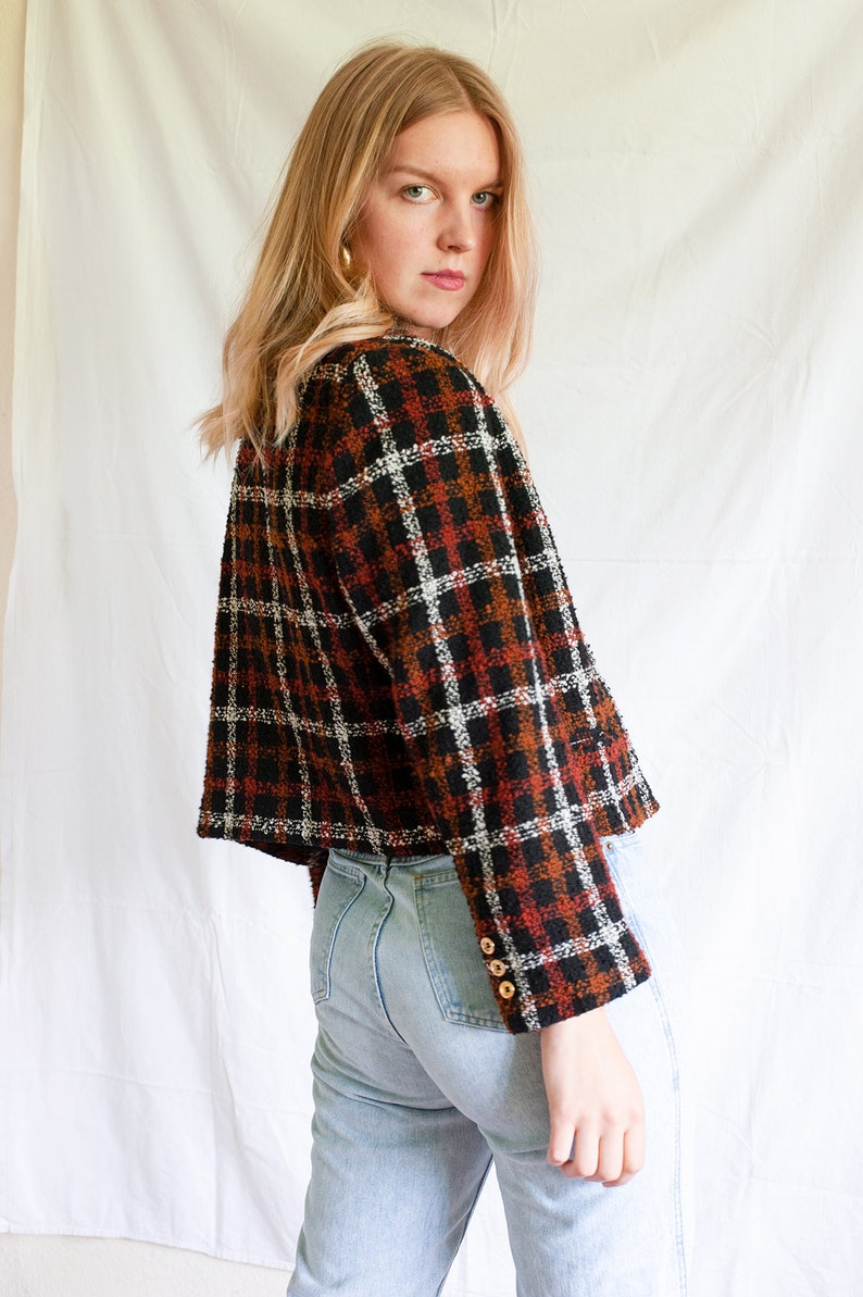 May include: A cropped plaid jacket with a black, white, and orange pattern. The jacket has long sleeves and a button closure. The model is wearing light blue jeans.