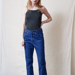 May include: A woman wearing a dark gray tank top and blue denim jeans with a wide leg.
