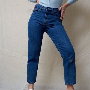 May include: A person wearing a light blue cardigan and dark blue jeans with a distressed detail on the left knee. They are wearing white heeled sandals.