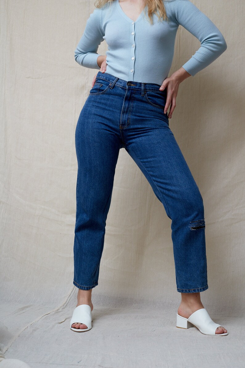 May include: A person wearing a light blue cardigan and dark blue jeans with a distressed detail on the left knee. They are wearing white heeled sandals.