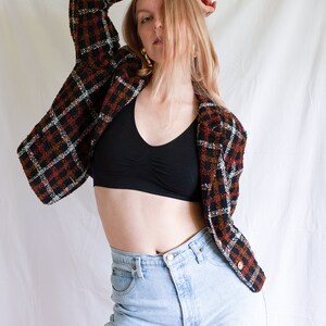 May include: A woman wearing a black crop top, light blue jeans, and a brown, black, and white plaid jacket.