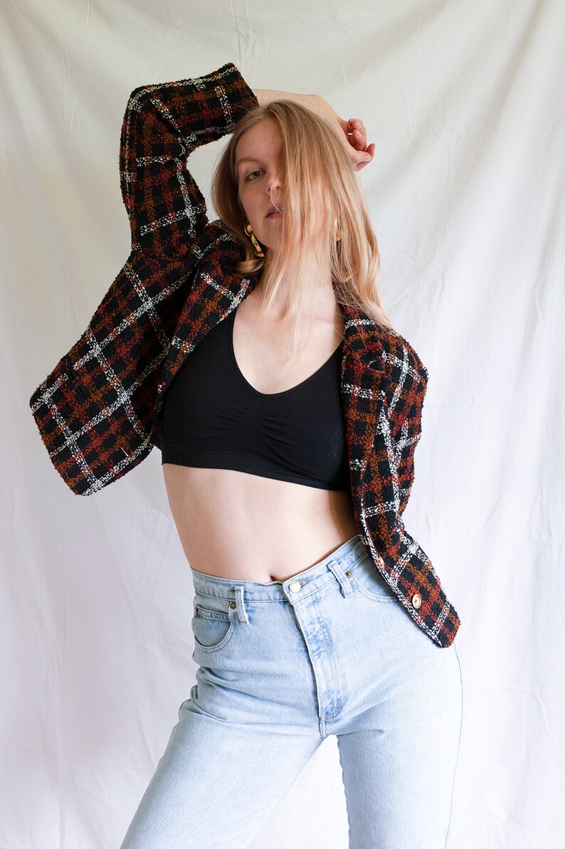May include: A woman wearing a black crop top, light blue jeans, and a brown, black, and white plaid jacket.