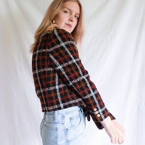 May include: A woman wearing a black, white, and orange plaid cropped jacket with gold buttons and light blue jeans.
