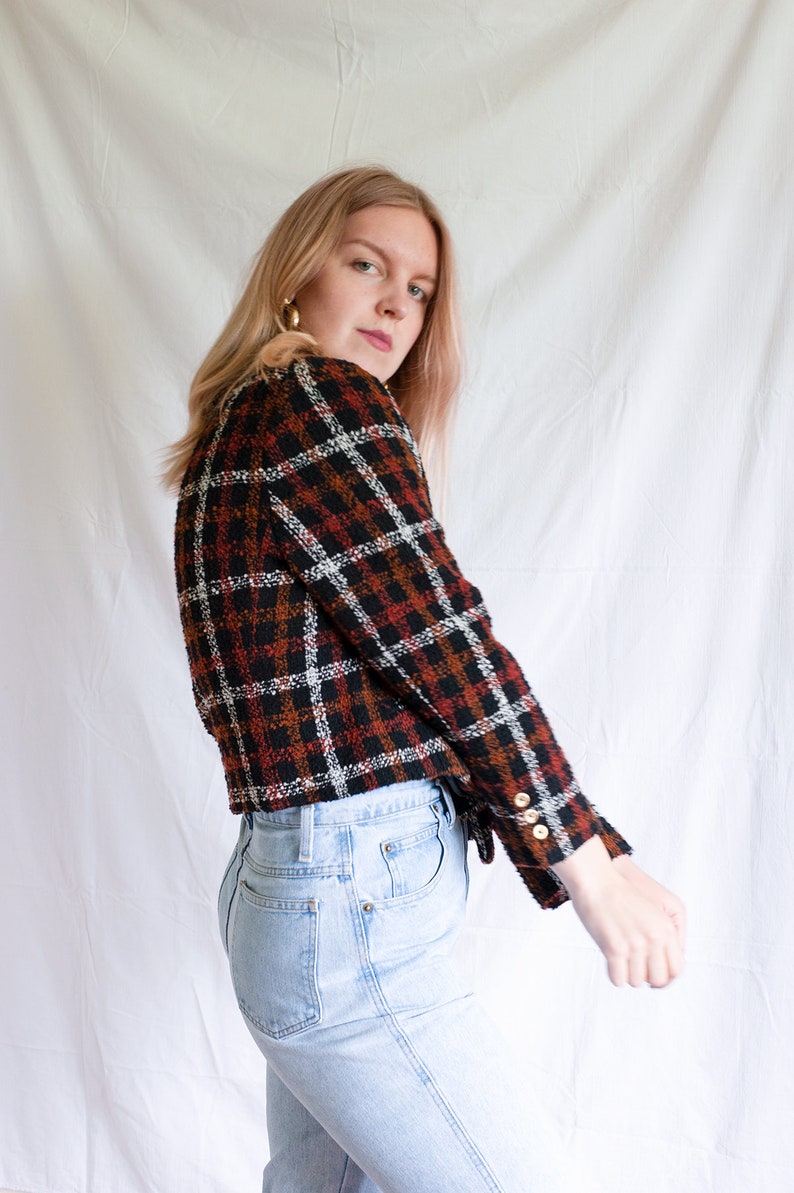 May include: A woman wearing a black, white, and orange plaid cropped jacket with gold buttons and light blue jeans.