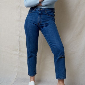 May include: A person wearing a light blue cardigan and dark blue jeans with a distressed knee. They are wearing white heeled sandals.