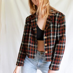May include: A cropped black, white, and orange plaid blazer with gold buttons. The blazer is worn over a black tank top and blue jeans.