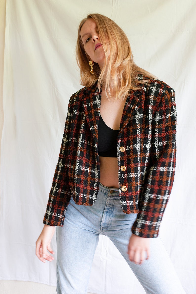 May include: A cropped black, white, and orange plaid blazer with gold buttons. The blazer is worn over a black tank top and blue jeans.