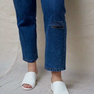 May include: A pair of white leather open-toe heeled sandals with a low block heel. The sandals are worn by a person wearing blue jeans with a distressed knee.