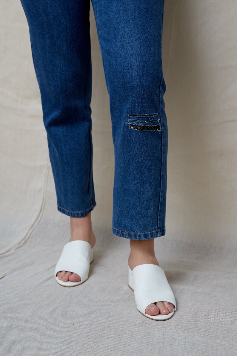 May include: A pair of white leather open-toe heeled sandals with a low block heel. The sandals are worn by a person wearing blue jeans with a distressed knee.