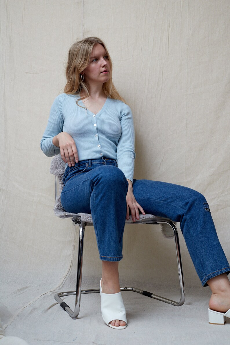 May include: A woman wearing a light blue cardigan and blue jeans sits on a chair. She is wearing white open-toe heeled shoes.