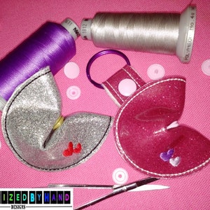 May include: Two fortune cookie-shaped keychains, one silver with red hearts and one pink with purple hearts. The keychains are on a pink surface with spools of thread and pink buttons.
