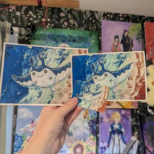 May include: Two art prints featuring a cartoon manta ray design with blue, white, and brown colors. The manta ray has a smiling face and is set against a swirling background. The prints are held up in front of other paintings.