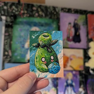 May include: A small, rectangular painting featuring a whimsical green creature with a round body, large eyes, and a blueberry. The background has blue and white swirls. The artwork is held by a hand.