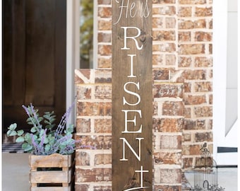 May include: A wooden sign with the words "He is Risen" in white lettering. The sign also includes a cross and the scripture reference "Matthew 28:6".
