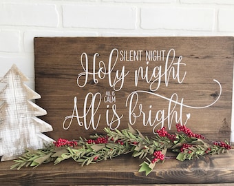 May include: A rustic wooden sign with white lettering that reads "Silent Night Holy night All is calm All is bright". The sign is decorated with a green garland with red berries.