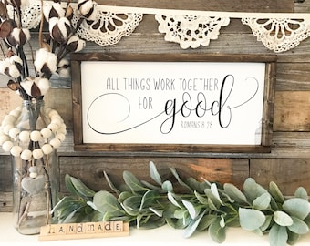 May include: A wooden sign with the text "All things work together for good, Romans 8:28" in black script font. The sign is framed with a dark brown wood frame and is sitting on a wooden surface with a green leafy garland and a glass jar with white beads and cotton stems.