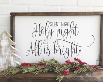May include: White wooden sign with black calligraphy that reads "Silent Night Holy Night All is Calm All is Bright". The sign is framed in dark wood and is decorated with a white wooden Christmas tree and a garland of green leaves and red berries.