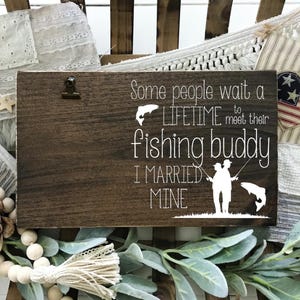 May include: A rustic wooden sign with the text "Some people wait a LIFETIME to meet their fishing buddy I MARRIED MINE." The sign features a clip and a white graphic of two people fishing. Decorative elements surround the sign.