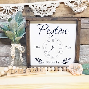 May include: A white wooden sign with a brown frame, featuring the name "Peyton" in black cursive script. A clock face with Roman numerals displays the time of birth, along with the weight and length of the baby. The sign also includes the date "04.30.18" and two leafy branches.