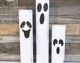 Puede incluir: Tres decoraciones de fantasmas de madera blanca con caras pintadas de negro. Los fantasmas son de diferentes tamaños y tienen diferentes expresiones faciales. El fantasma más grande tiene dos ojos y una boca, el fantasma del medio tiene dos ojos y una amplia sonrisa, y el fantasma más pequeño tiene dos ojos y una boca pequeña.