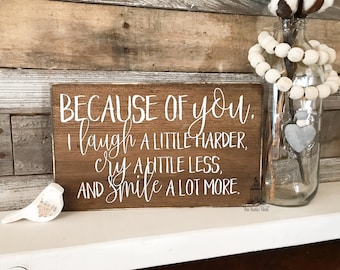 Könnte beinhalten: Ein Holzschild mit weißer Aufschrift, die lautet: "BECAUSE OF you. I laugh A LITTLE HARDER, cry A LITTLE LESS, AND smile A LOT MORE."